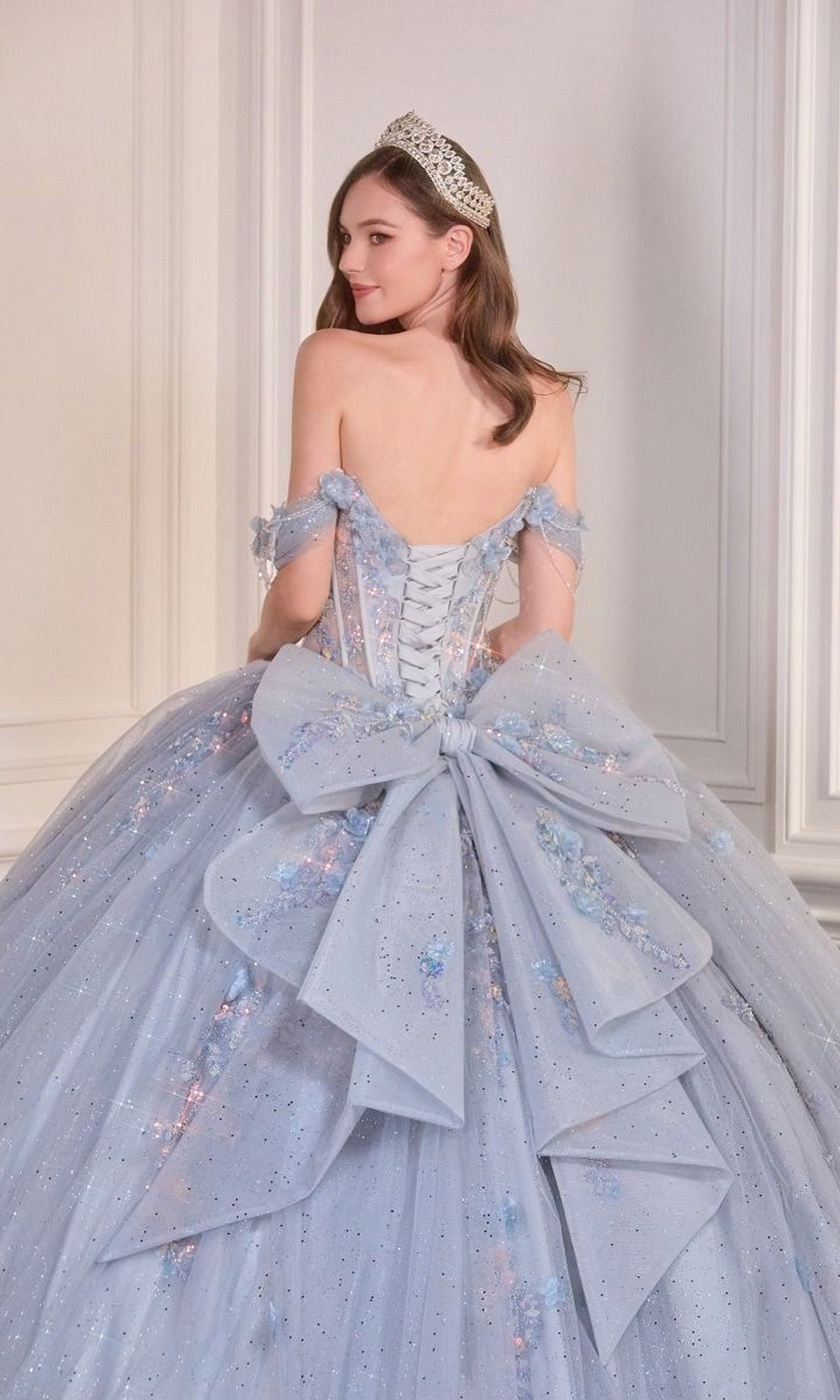 A woman in the Quinceanera Dress 15728 By Ladivine, featuring sparkle tulle, a large back bow, and a tiara, stands before a white paneled wall, glancing over her shoulder.